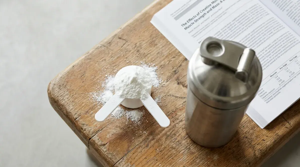 A measured 5-gram scoop of white creatine monohydrate powder beside a stainless steel shaker bottle on a gym bench, with an open peer-reviewed journal article on creatine timing in the background, soft daylight